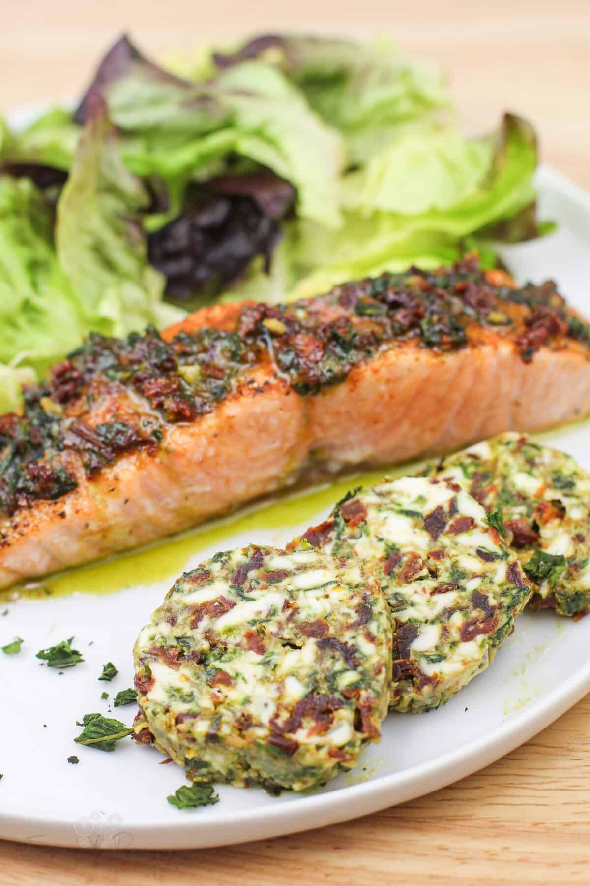 plated salmon and salad on white plate; sliced sundried tomato basil finishing butter in front
