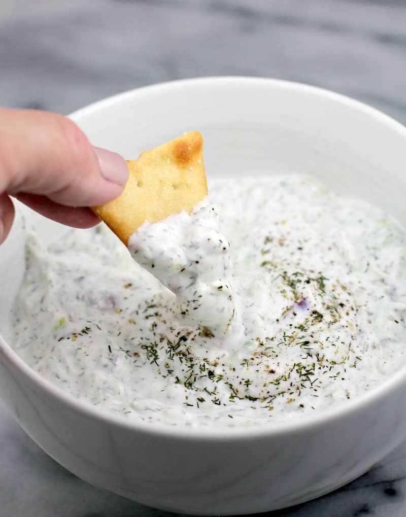 a hand scooping Greek yogurt dip tzatziki sauce with a chip in a white bowl with marble background