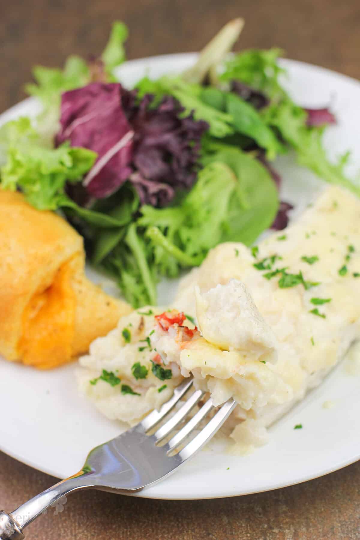 folk holding bite of prepared haddock with lobster sauce on white plate with salad and roll