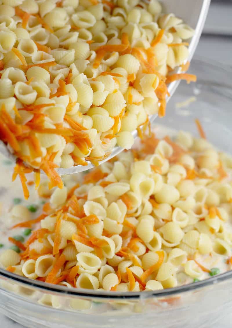 cooked pasta shells and shredded carrot being added to the remaining ingredients in a glass bowl