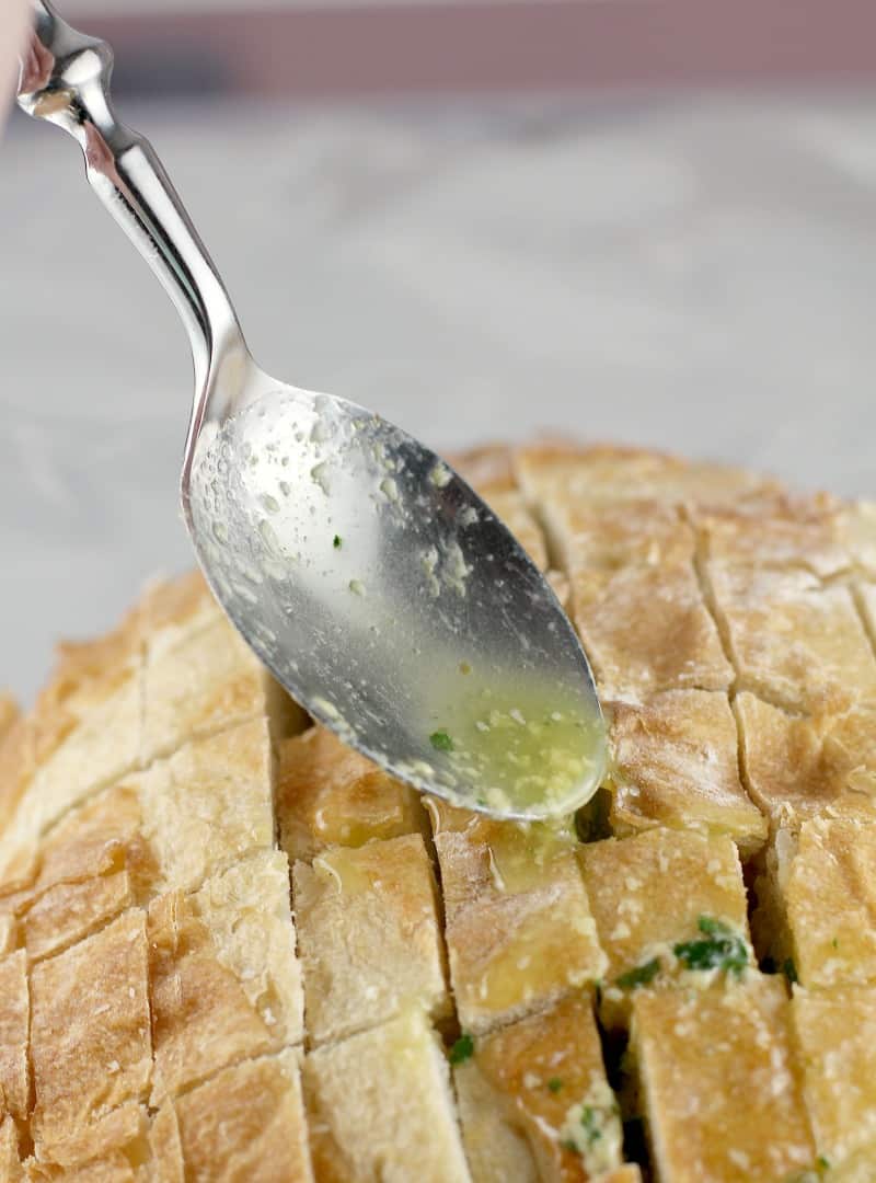 a spoon drizzling in the butter herb mixture into cut cracks on the bread loaf