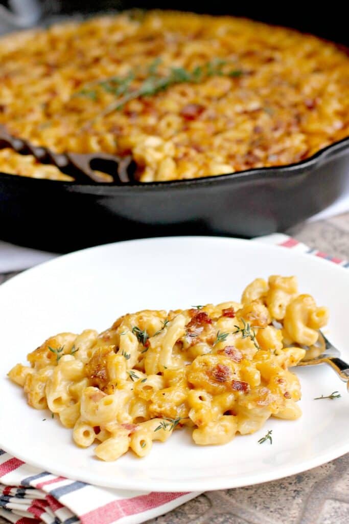 side view of finished bacon macaroni and cheese plated on a white dish; remainder of the mac n cheese is in the background in a cast iron skillet