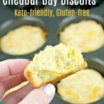hand holding biscuit broken in half to show flaky inside, other biscuits in muffin tin in the background, pinnable image